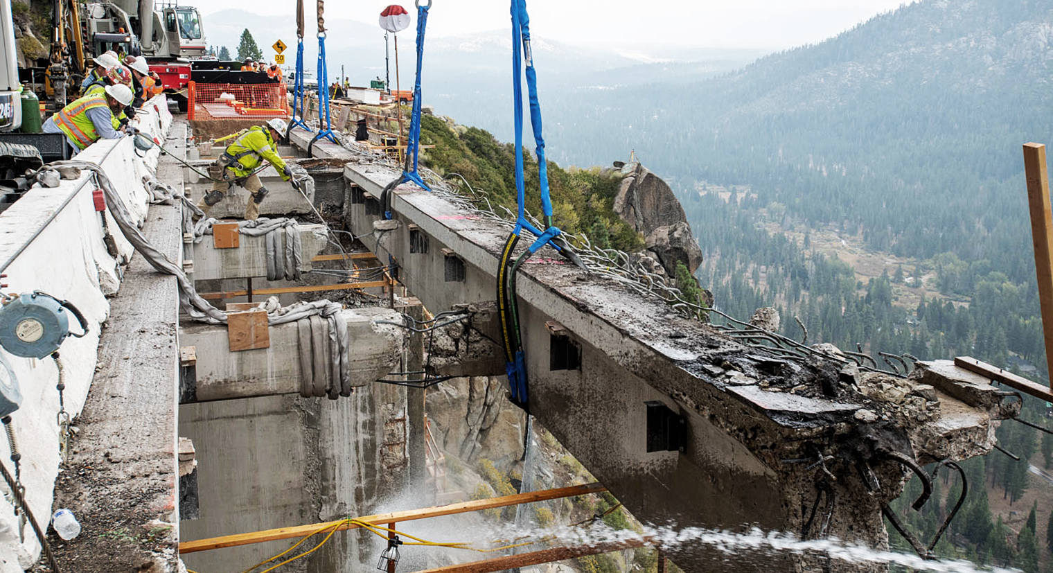 Photo of workers hoisting old bridge guardrail. Mountain scenery in background.