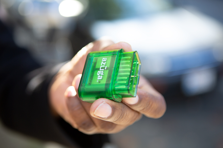 Photo close up of a hand holding the Azuga device with a car in the background.